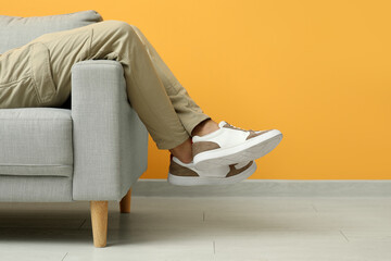 Wall Mural - Young man in stylish sneakers sitting on sofa near yellow wall, closeup