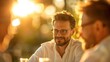 © KanitChurem - Smiling man with glasses enjoying an outdoor gathering with friends during sunset, creating a warm and cheerful atmosphere.