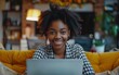 © imagineRbc - Cheerful black woman with curly hair working at home on laptop, smiling. Sitting by window with green plant in foreground, garden view outside