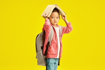 Wall Mural - Cute chinese elementary school girl holding book above head and having backpack on shoulders, standing on yellow isolated background. Asian ethnicity six year old kid preparing to get school. 