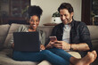© peopleimages.com - Couple, relax and happy with technology on couch for social media, streaming subscription and online shopping for apartment. Man, woman and together in home with laptop or smartphone for connectivity
