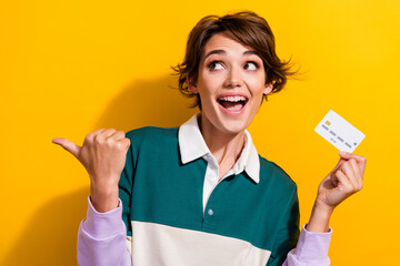 Poster - Portrait of pretty nice young girl hold debit card indicate finger empty space wear shirt isolated on yellow color background