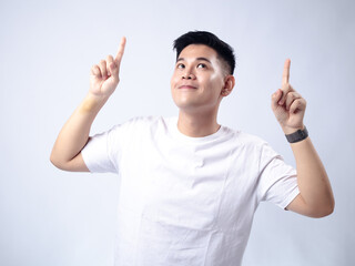 A young Asian man, wearing a white shirt and black watch, looks up while pointing both index fingers upwards. The background is plain white