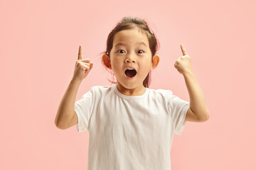 Wall Mural - Sweet korean child girl looks amazed points at something up and looking to camera, isolated on a pink background. Cute little kid sees an amazing thing up, gestures indoor