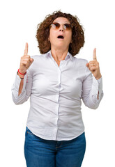 Poster - Beautiful middle ager senior business woman wearing sunglasses over isolated background amazed and surprised looking up and pointing with fingers and raised arms.