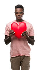 Poster - Young african american man holding red heart with a confident expression on smart face thinking serious