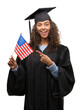 © Krakenimages.com - Young hispanic woman wearing graduation uniform holding flag of United States very happy pointing with hand and finger