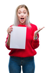 Wall Mural - Young blonde woman holding blank paper sheet over isolated background very happy and excited, winner expression celebrating victory screaming with big smile and raised hands