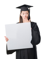 Wall Mural - Young Chinese woman wearing graduate uniform holding banner cover mouth with hand shocked with shame for mistake, expression of fear, scared in silence, secret concept