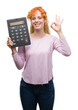 © Krakenimages.com - Young redhead woman holding big calculator doing ok sign with fingers, excellent symbol