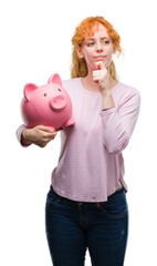 Wall Mural - Young redhead woman holding piggy bank serious face thinking about question, very confused idea