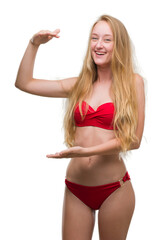 Canvas Print - Blonde teenager woman wearing red bikini gesturing with hands showing big and large size sign, measure symbol. Smiling looking at the camera. Measuring concept.