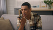© Yuliia - Happy African American man at home couch sofa biracial male ethnic guy using cellphone answering on audio message record voice mail recording answer in social media voicemail use speakerphone aloud