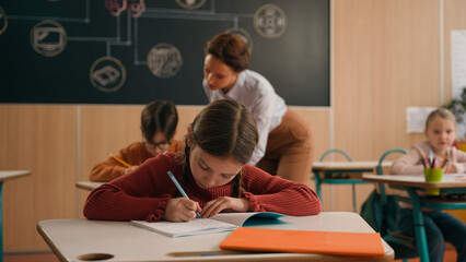 School class lesson children kids studying learning woman teacher female walking teaching little boy child kid rise hand ask help tutor check task study learn helping write talking primary education