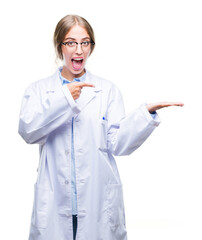 Canvas Print - Beautiful young blonde doctor woman wearing white coat over isolated background amazed and smiling to the camera while presenting with hand and pointing with finger.