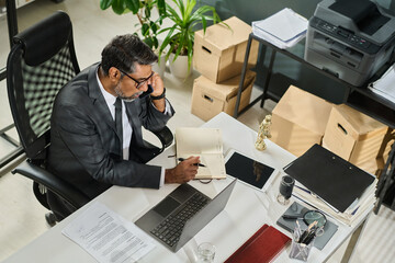 Wall Mural - High angle of mature male ceo or director of financial company sitting by workplace and speaking on mobile phone while organizing work