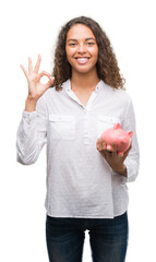 Sticker - Young hispanic woman holding piggy bank doing ok sign with fingers, excellent symbol