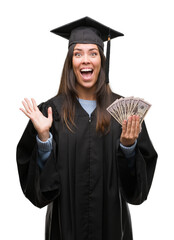 Wall Mural - Young hispanic woman wearing graduated uniform holding dollars very happy and excited, winner expression celebrating victory screaming with big smile and raised hands