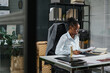 © pressmaster - Young serious woman in eyeglasses and white shirt sitting by workplace in office and looking through business contract before signing