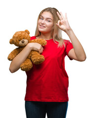 Poster - Young caucasian woman holding cute teddy bear over isolated background doing ok sign with fingers, excellent symbol