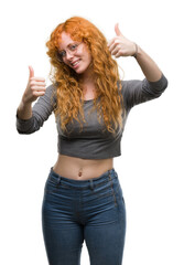 Wall Mural - Young redhead woman approving doing positive gesture with hand, thumbs up smiling and happy for success. Looking at the camera, winner gesture.