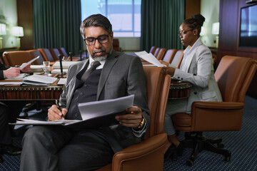 Wall Mural - Serious male chief executive officer looking through new business contract before signing it while sitting in armchair against colleagues