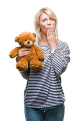 Poster - Young beautiful blonde woman holding teddy bear plush over isolated background cover mouth with hand shocked with shame for mistake, expression of fear, scared in silence, secret concept