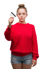 Poster - Young blonde woman holding credit card with a confident expression on smart face thinking serious