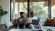 © Oscar - An LGBT couple sitting on a modern sofa, both engaged with their devices. The bright and airy room features large windows and lush greenery outside.