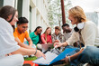 © Jose Calsina - Group of multiracial students doing homework together, sitting on the university campus lawn. Teenagers sharing notes to study for their high school exam. Young people talking and learning the lesson