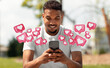 © Prostock-studio - A young black man is sitting in a park, smiling while looking at his smartphone. He is holding the phone with both hands, and there are multiple heart-shaped icons floating around him