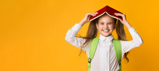 Wall Mural - Home School Reading. Positive Elementary Schoolgirl Holding Book On Head Smiling To Camera Over Yellow Background. Studio Shot