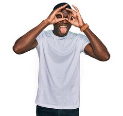 Wall Mural - Young african american man wearing casual white t shirt doing ok gesture like binoculars sticking tongue out, eyes looking through fingers. crazy expression.