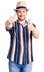 Wall Mural - Young handsome caucasian man wearing summer hat approving doing positive gesture with hand, thumbs up smiling and happy for success. winner gesture.