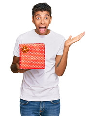 Poster - Young handsome african american man holding gift celebrating victory with happy smile and winner expression with raised hands