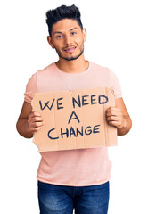 Wall Mural - Handsome latin american young man holding we need a change banner looking positive and happy standing and smiling with a confident smile showing teeth