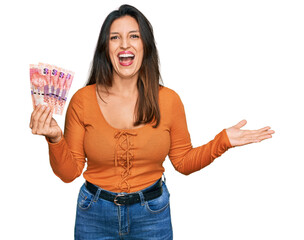 Wall Mural - Beautiful hispanic woman holding south african 50 rand banknotes celebrating victory with happy smile and winner expression with raised hands