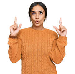 Wall Mural - Young brunette woman wearing casual winter sweater amazed and surprised looking up and pointing with fingers and raised arms.