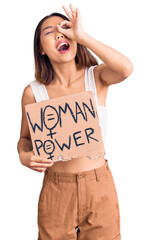 Young beautiful chinese girl holding woman power banner smiling happy doing ok sign with hand on eye looking through fingers