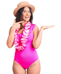 Wall Mural - Young beautiful brunette woman wearing swimwear and hawaiian lei amazed and smiling to the camera while presenting with hand and pointing with finger.