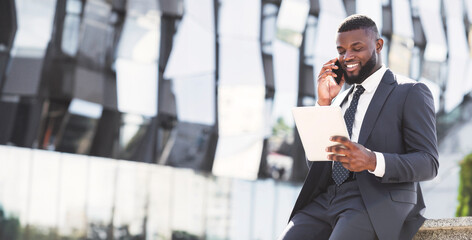 Wall Mural - Positive African Entrepreneur Talking On Cellphone Using Digital Tablet During Coffee Break In City Urban Area