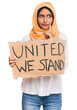 © Krakenimages.com - Young brunette arab woman holding united we stand banner serious face thinking about question with hand on chin, thoughtful about confusing idea