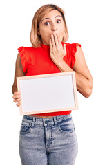 Poster - Young blonde woman holding empty white chalkboard covering mouth with hand, shocked and afraid for mistake. surprised expression