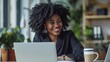 © Prostock-studio - A woman with curly hair smiles while working on a laptop at her desk. A coffee cup sits to the right of her, and plants are visible in the background.