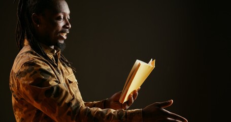 Wall Mural - Young adult reading literature book on camera, standing over black background and enjoying lecture as hobby. African american person having fun with fiction or action novel story. Handheld shot.