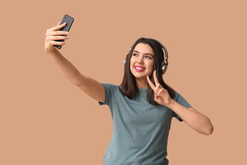 Wall Mural - Beautiful young woman in modern headphones taking selfie and showing victory gesture on brown background