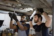 © sofiko14 - Two mechanics performing repair on car undercarriage in garage workshop. Focus on teamwork, dedication, and technical skill. Professional automotive maintenance environment.