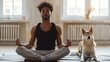 © liliyabatyrova - A young African American man in a black tank top meditates in a yoga pose on a mat, eyes closed, while his dog sits nearby
