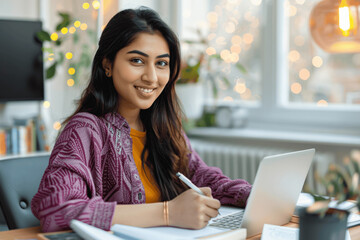 Wall Mural - Smiling young Indian woman taking notes on laptop at home office engaged in online learning and virtual meetings for work and education