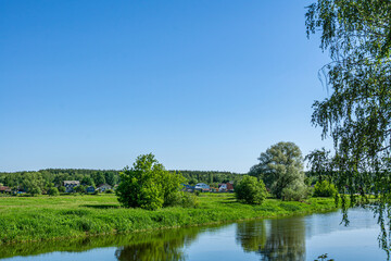 Wall Mural - Summer countryside landscape with village and river under blue sky. Rural river with green meadow peaceful landscape.